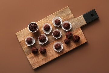 Wooden board with delicious chocolate truffles in paper cups and bowl of chocolate paste on brown background