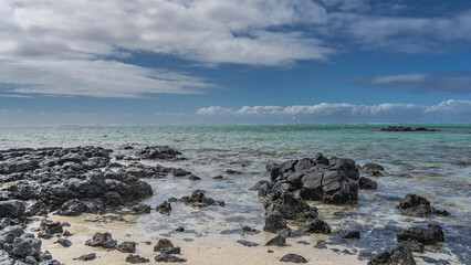 The endless turquoise ocean. Black volcanic boulders are scattered on the sandy beach near the shore. Crystal clear water. Blue sky, clouds. Mauritius.