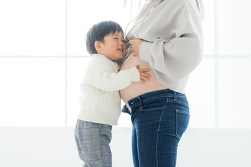 屋内の自然光豊かなスタジオで臨月のお母さんのお腹に抱きついて幸せそうな表情をする子どものマタニティフォト