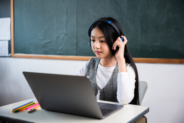 A girl is sitting at a desk with a laptop in front of her. She is wearing headphones and she is...