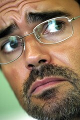 A close-up of a man with glasses displaying a thoughtful expression and intense gaze, taken in a neutral setting during daylight hours
