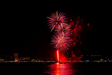 Brilliant red fireworks explode against a dark night sky, illuminating a cityscape below. Vibrant...
