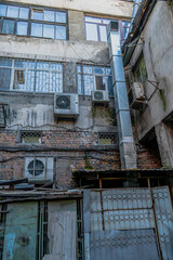 An exterior view of a weathered industrial building with visible air conditioning units, ductwork, and iron grills on windows. The structure showcases rugged textures, aged brickwork, and urban decay 