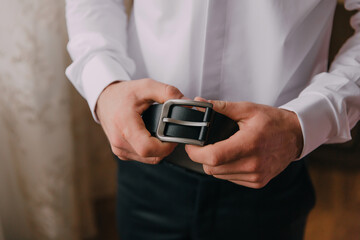 A man is wearing a belt and holding it up to his waist. The belt is black and has a silver buckle. The man is wearing a white shirt and black pants. Concept of formality and professionalism