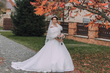 Fototapeta premium A bride is standing in front of a tree with a white dress and a fur stole. She is holding a bouquet of flowers