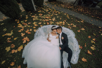 A bride and groom are lying on a bench covered in leaves. The bride is wearing a white dress and...