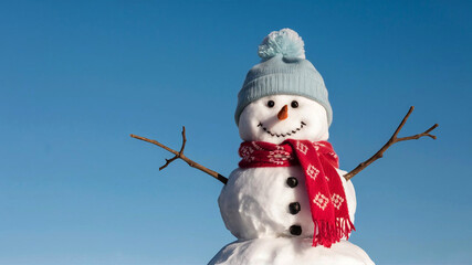 Obraz premium A charming snowman complete with a cozy red scarf and a fluffy blue hat smiles warmly as it stands against a bright blue background evoking the magic of winter.