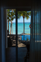 Breathtaking view from a luxury hotel room in Mauritius. Framed by soft, sheer curtains, the scene outside reveals a turquoise ocean stretching to the horizon with gentle waves. Palm trees sway nearby