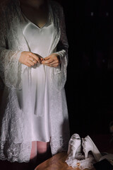 A woman in a white gown is getting ready for a wedding. She is holding a pair of white shoes and...