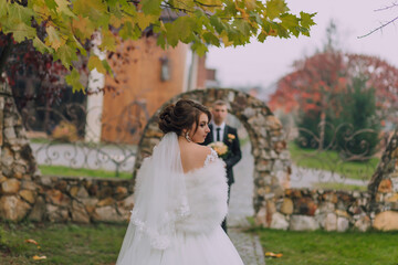 A bride and groom are standing in front of a stone archway. The bride is wearing a white dress and...