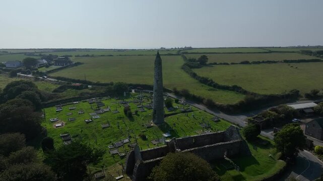 St. Declan's Monastery, Ardmore, County Waterford, Ireland, September 2024. Drone wide angle clockwise orbit around the Round Tower and graveyard separated by the leafy tree line on an overcast day.