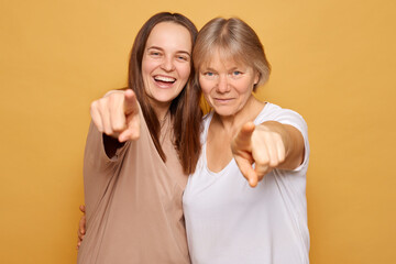 Two smiling women enthusiastically pose for the camera with their fingers pointed outward against a vibrant yellow backdrop