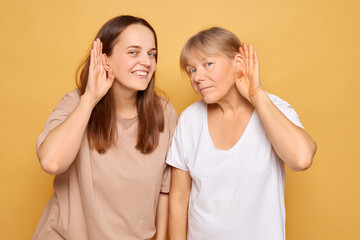 Two women standing against a yellow background, playfully cupping their ears and leaning slightly forward as if trying to listen closely during a lighthearted moment