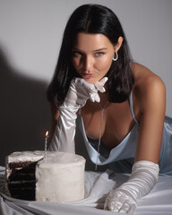 An elegant woman in a light blue satin dress and white gloves leans over a birthday cake with lit...