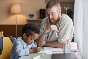 Medium shot of happy supportive father helping little adopted son with homework while sitting at...