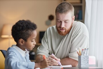 Medium shot of supportive dad helping little adopted son with homework while sitting at desk and...
