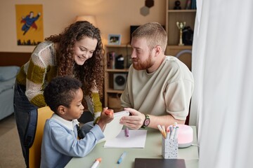Medium shot of happy adoptive mother and father engaged in hand drawing with little Black boy...