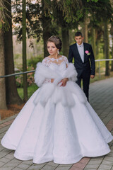 A bride and groom are walking down a path in a forest. The bride is wearing a white dress and a fur stole, while the groom is wearing a suit. The scene is romantic and picturesque