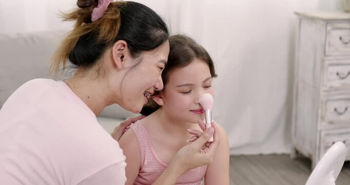 Asian mother affectionately applies blush to young daughter's nose, both sharing a tender smile. depicts love, bonding, and warmth in a relaxed home environment, showcasing gentle family interactions.