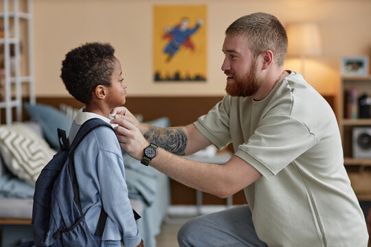 Side view of happy adoptive father adjusting sons clothes while preparing little Black boy for school in kids room