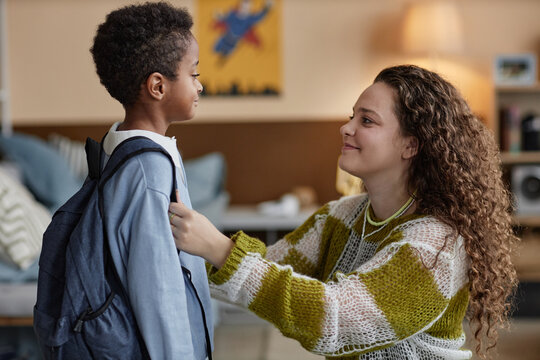 Side view of happy adoptive mother adjusting sons clothes while preparing little Black boy for school in kids room