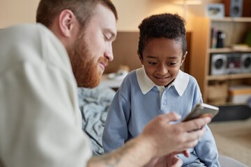 Medium shot of smiling little Black boy looking at screen while dad showing funny video on phone in...