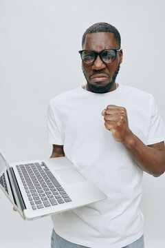 frustrated young man with laptop, expressing anger, wearing glasses, white t shirt, gray background, showing emotions of disappointment and irritation while working on the computer