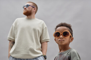 Medium shot of stylish little Black boy in large sunglasses looking away with adoptive father standing in background wearing resembling accessory against gray background in studio, copy space