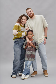 Vertical full length shot of happy adoptive parents posing in gentle hug with little Black boy while standing against gray background in studio