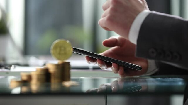 A man is holding a cell phone and a stack of coins on a table
