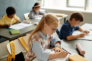 Fototapeta premium Side view of young blonde girl in glasses diligently writing in notebook while sitting at desk during lesson in classroom, copy space