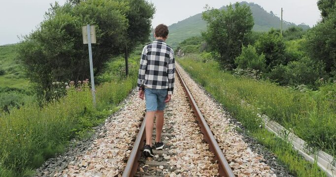 Teenager in casual plaid shirt and shorts walking along train tracks in a rural landscape, filmed from behind, portraying teenage loneliness and isolation