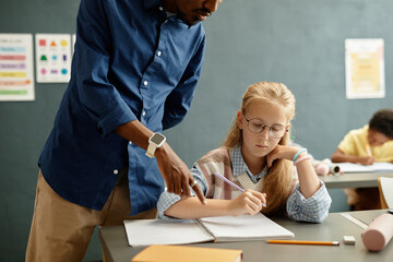 Young blonde girl with glasses sitting at desk writing test in class while male teacher helping student pointing at copybook making remark at school, copy space