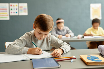 Medium shot of young boy focused on writing task in copybook sitting at desk while studying in classroom with blue wall at school, copy space