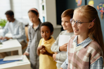 Fototapeta premium Side view of smiling young girl wearing glasses standing in line with classmates talking in front of class during lesson in primary school classroom, copy space