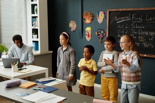 Adorable young students from diverse ethnic backgrounds standing in front of class presenting results of collaborative task to classmates during lesson in modern classroom