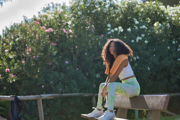 Naklejka premium Latina woman, young, beautiful, brunette, with curly hair, wearing sports clothes, headphones and towel, sitting on a bench relaxed and calm after exercise. Concept sport, exercise, rest, active.