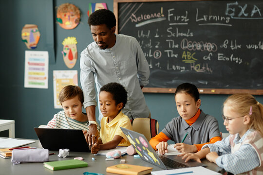Diverse group of primary school children using laptops in pairs to study sitting at large desk in classroom while African American male teacher helping kids working on computers, copy space