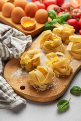 Uncooked tagliatelle pasta nests with basil, tomatoes and eggs on wooden cutting board
