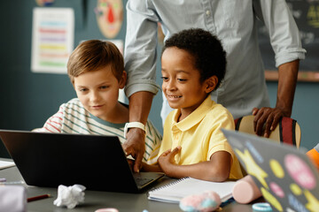 Medium shot of young African American boy with adorable smile using laptop with classmate watching educational video, while unrecognizable teacher helping kids working on computer in class