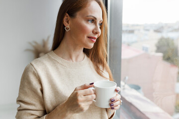 Portrait of beautiful well-groomed elegant woman entrepreneur standing with cup of coffee in hands looking through window at city in morning before going for work. Calm peaceful life