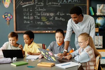 Modern male teacher of African American ethnicity helping young girls with working on laptop in...