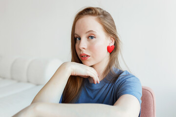 Portrait of puzzled pensive concerned and thoughtful redhead teen girl with heart-shaped red earrings sitting on chair in her white cozy room, thinking of boys and its behavior, touching her chin