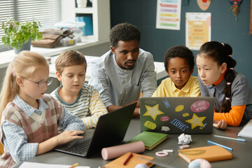 Supportive African American male teacher helping children use laptops sitting together with kids at large table, while explaining homework assignment in modern classroom