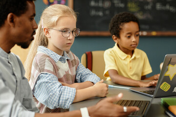 Side view of young blonde girl sitting at desk with laptop listening attentively to teachers instructions on how to complete homework assignment using laptop in modern classroom, copy space