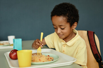 Medium shot of young African American boy sitting at table enjoying eating tasty lunch in school cafeteria, copy space
