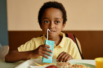 Medium shot of young African American boy drinking fresh juice or milk from straw while enjoying lunch in school cafeteria, copy space