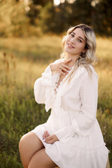 Beautiful Caucasian blonde woman sits on a wooden chair in a field. Model poses in a white dress in nature. Concept of natural and organic beauty and youth.