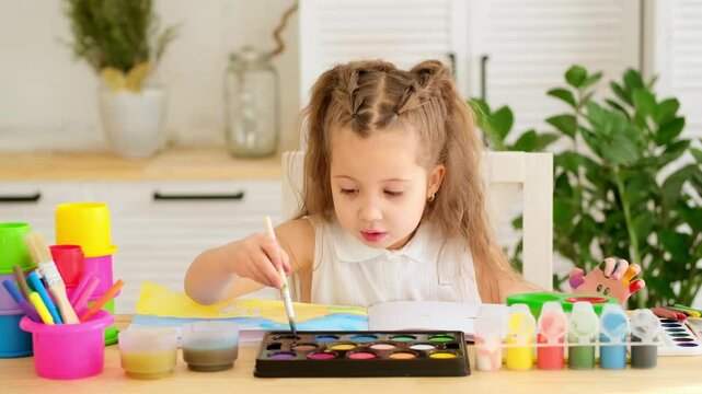 a happy and joyful child paints at a table in a bright room, a little girl learns to draw with a brush and gouache, a drawing lesson