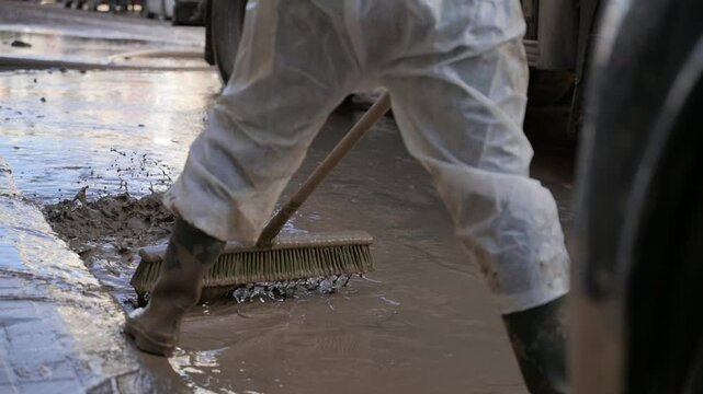 A person in protective gear sweeps a muddy street with a large brush, following a major flood in Valencia